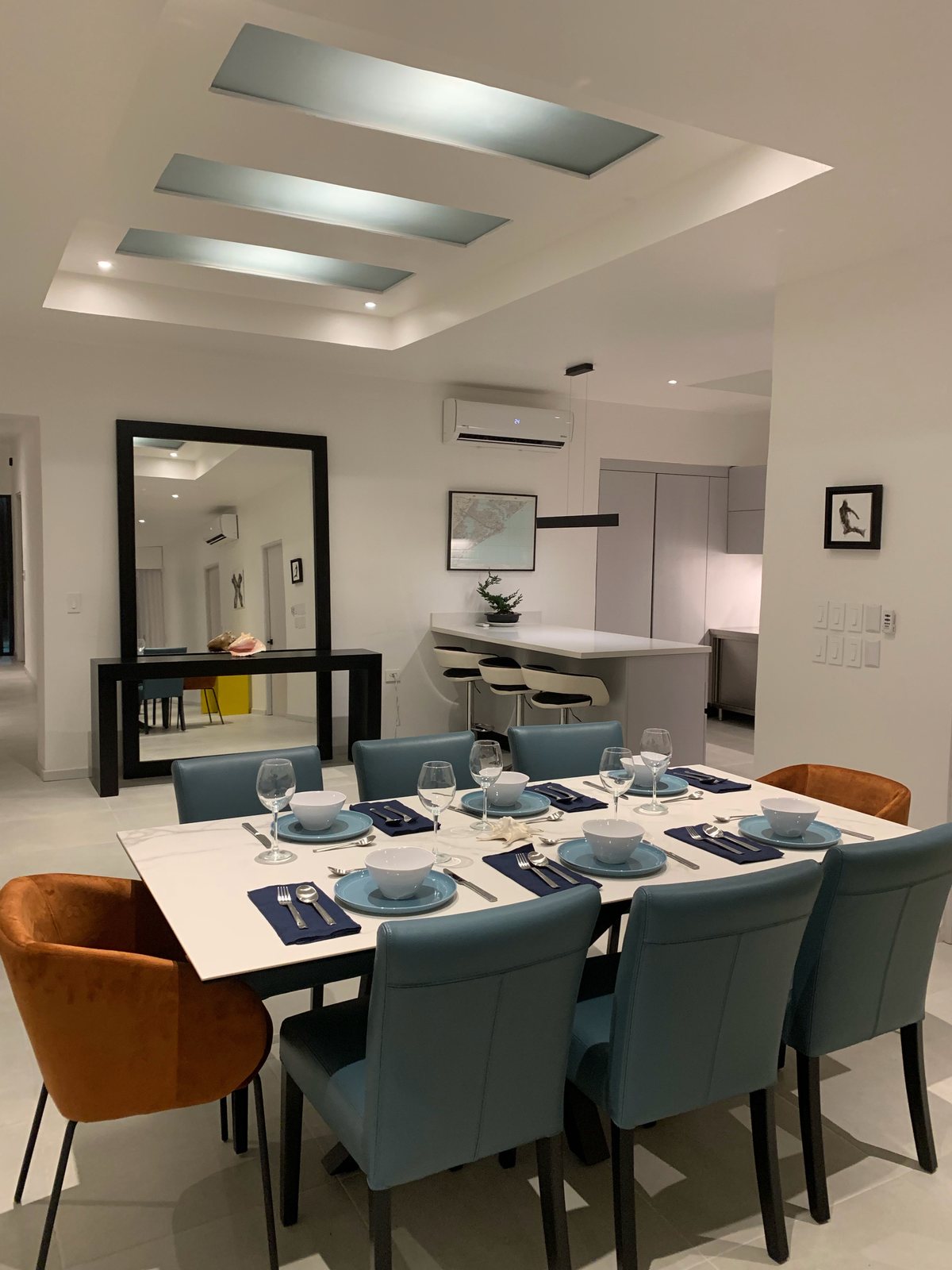 Dining room with a white table set for eight, blue leather chairs and orange armchairs, with the kitchen bar behind